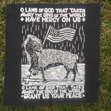 black canvas patch with text O LAMB OF GOD THAT TAKES AWAY THE SINS OF THE WORLD HAVE MERCY ON US O LAMB OF GOD THAT TAKES AWAY THE SINS OF THE WORLD GRANT US YOUR PEACE with a drooling wolf draped in the skin of a dead lamb stomping on a cross and holding an American flag in a pose reminiscent of the classic Christian iconography of a lamb holding a flag with a cross.